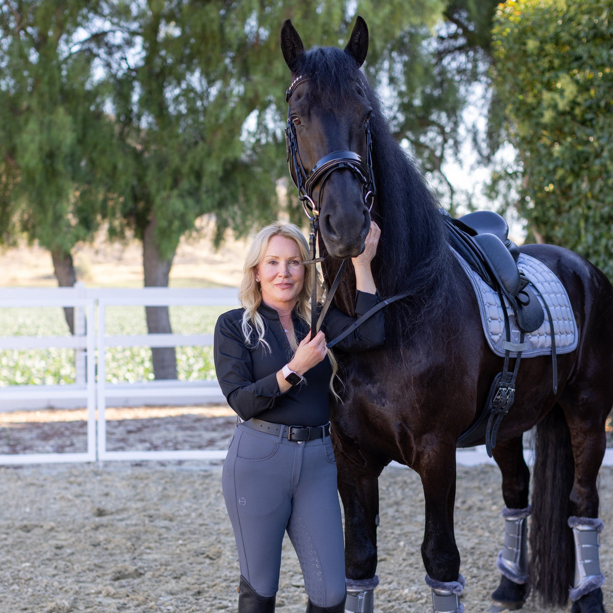 Woman in equestrian attire standing next to a dark horse in an outdoor setting with trees and a fence.