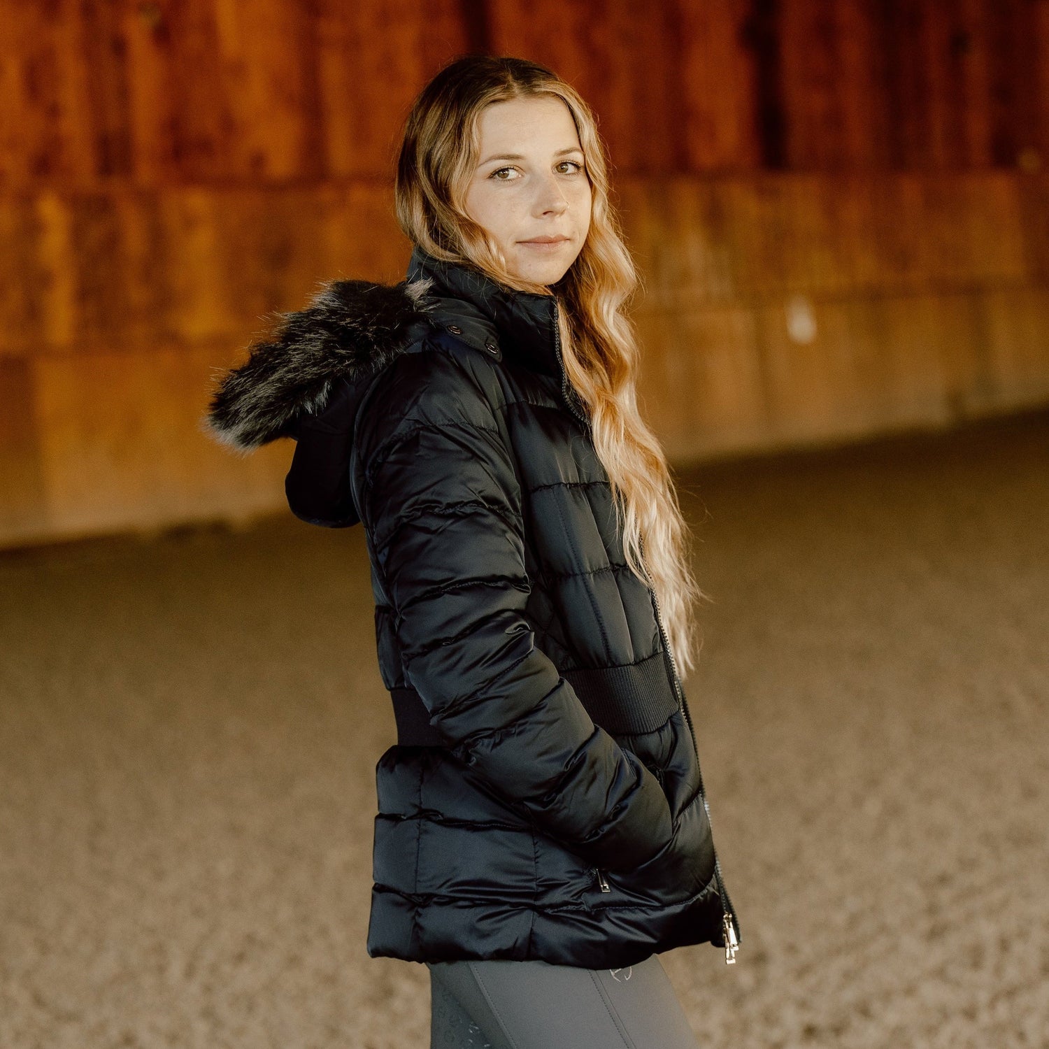 Person wearing a black puffer jacket and gray leggings standing in an indoor equestrian arena.
