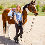 A woman standing next to a brown and white horse, wearing a sky blue short sleeve competition shirt with lace detailing and a metallic embroidered logo on the back.
