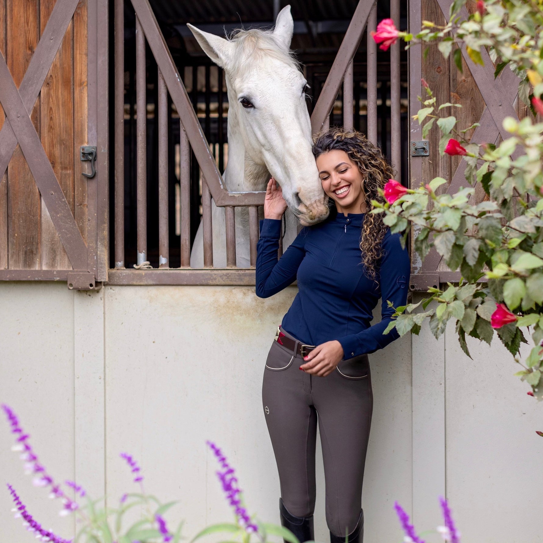 Woman in equestrian attire standing next to a horse in front of a stable.
